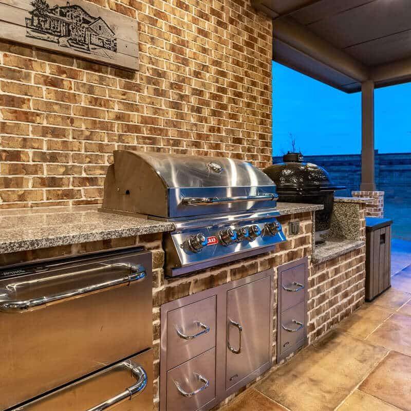 Brick outdoor kitchen with granite counters, a built-in grill and a kamado smoker under a covered patio